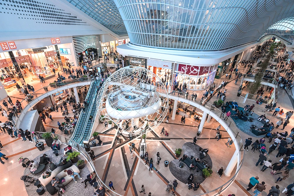 Aerial view of Chadstone shopping centre, surrounded by multiple levels of retail stores, escalators, and large crowds of shoppers throughout.