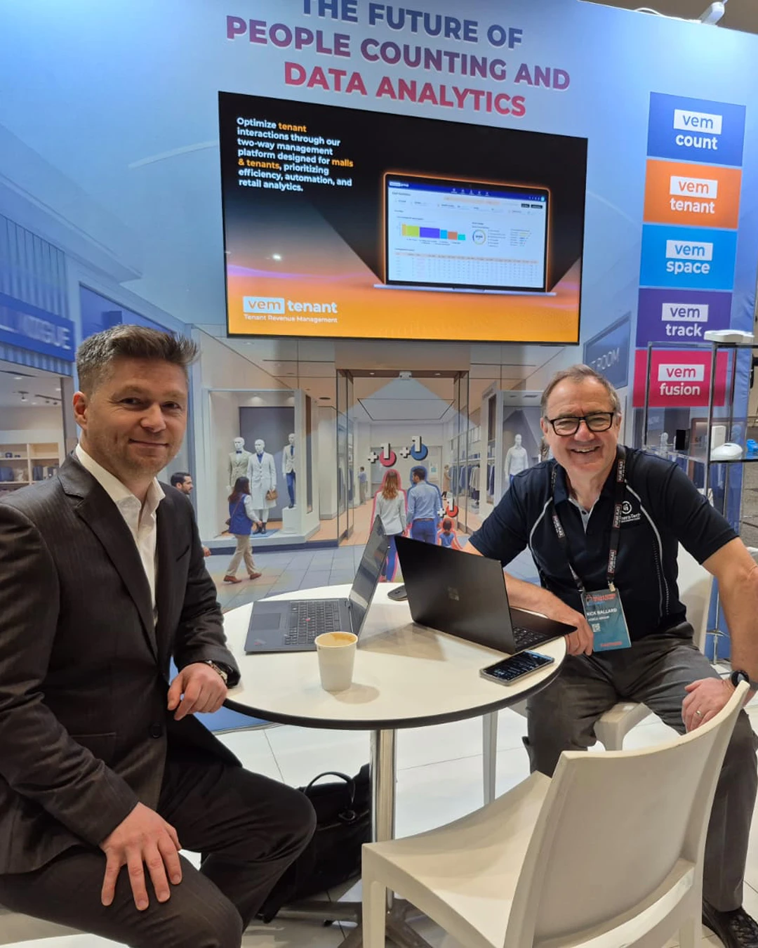 Two team members at the Cohera Tech exhibition stand at the Malls of the Future conference, with a large display screen showing "The Future of People Counting and Data Analytics".