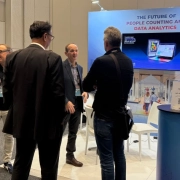 Two attendees in conversation on the exhibition floor at a retail technology conference, with a large branded display screen visible in the background.