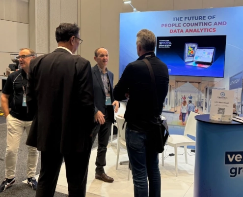 Two attendees in conversation on the exhibition floor at a retail technology conference, with a large branded display screen visible in the background.