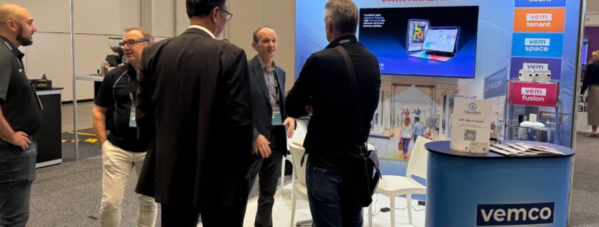 Two attendees in conversation on the exhibition floor at a retail technology conference, with a large branded display screen visible in the background.