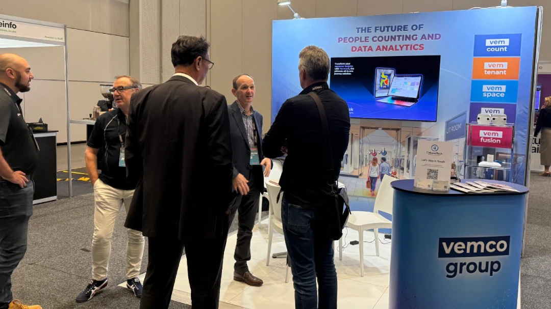 Two attendees in conversation on the exhibition floor at a retail technology conference, with a large branded display screen visible in the background.